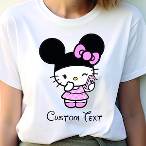 Pu&ograve; includere: T-shirt bianca con un personaggio dei cartoni animati con orecchie nere, un fiocco rosa e un vestito rosa, che tiene in mano un telefono. Il testo "Custom Text" &egrave; scritto in corsivo sotto.