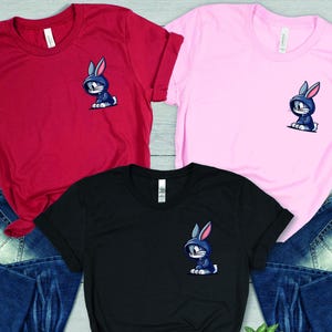 May include: Three t-shirts in red, pink, and black with a cartoon image of a rabbit wearing a blue hoodie on the front. The shirts are laid out on a wooden surface with blue jeans.