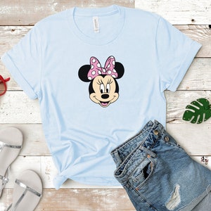 May include: A light blue t-shirt with a cartoon image of Minnie Mouse wearing a pink bow with white polka dots. The t-shirt is laid out on a wooden surface with a pair of silver sandals, a pair of red sunglasses, and a pair of blue denim shorts.