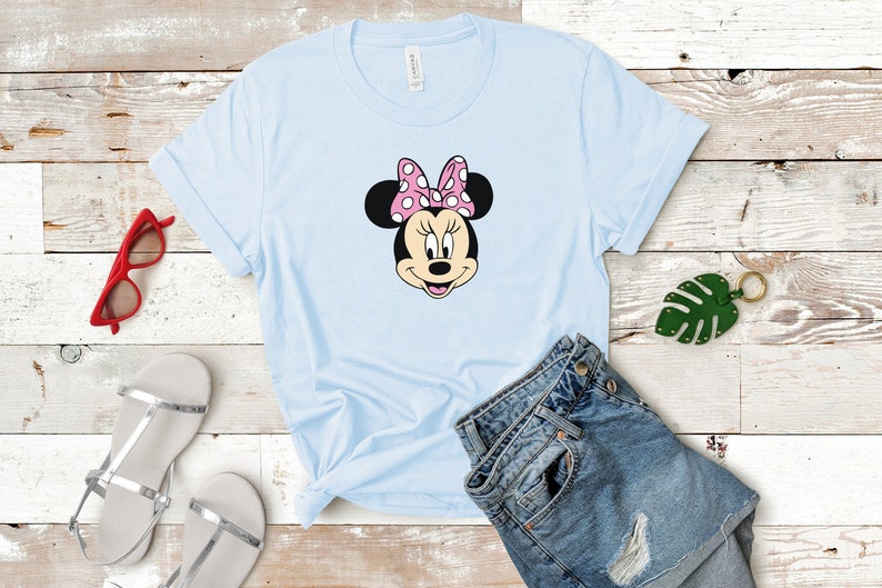 May include: A light blue t-shirt with a cartoon image of Minnie Mouse wearing a pink bow with white polka dots. The t-shirt is laid out on a wooden surface with a pair of silver sandals, a pair of red sunglasses, and a pair of blue denim shorts.