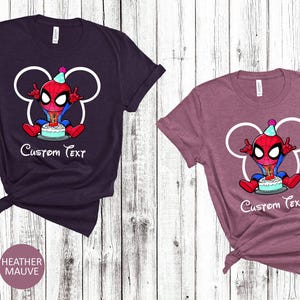 May include: Two t-shirts, one heather maroon and one heather mauve, featuring a cartoon Spider-Man character with a birthday cake. The design includes a white outline of Mickey Mouse ears and the text "Custom Text".