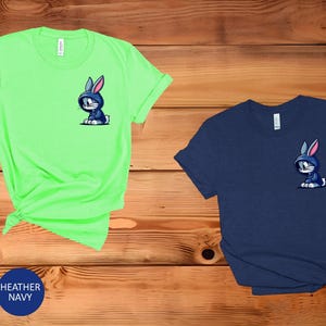 May include: Two t-shirts with a cartoon rabbit wearing a blue hoodie. The t-shirt on the left is a bright green color and the t-shirt on the right is a dark blue color. The text "MINT" and "HEATHER NAVY" is displayed below the t-shirts.