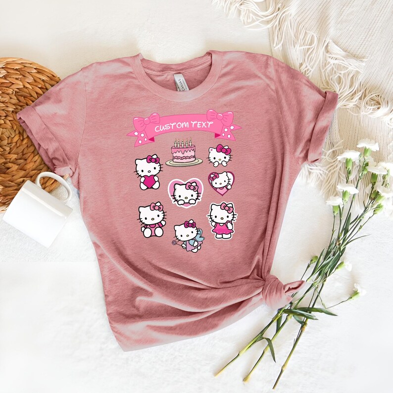 May include: A dusty rose-colored t-shirt featuring a collection of Hello Kitty graphics. The design includes various poses of the character, a cake, and hearts. A banner at the top reads "CUSTOM TEXT" in pink. The shirt is styled with rolled-up sleeves.