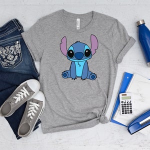 May include: A gray t-shirt with a blue cartoon character on the front. The character has large eyes, a blue body, and pink ears. The shirt is laid out on a white wooden surface with a pair of jeans, a water bottle, a calculator, a stapler, and a notebook.