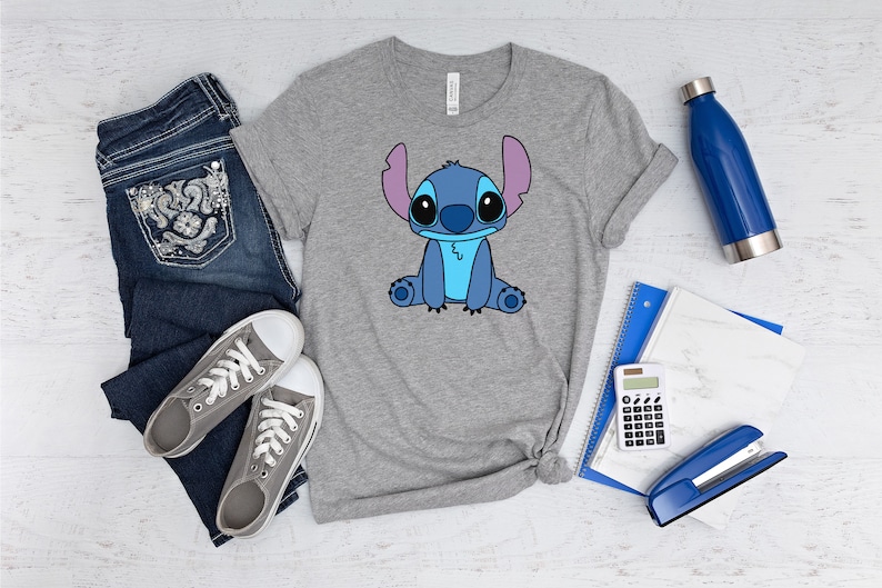 May include: A gray t-shirt with a blue cartoon character on the front. The character has large eyes, a blue body, and pink ears. The shirt is laid out on a white wooden surface with a pair of jeans, a water bottle, a calculator, a stapler, and a notebook.