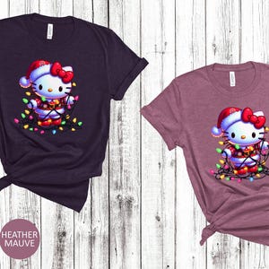 May include: Two t-shirts, one heather maroon and one heather mauve, each featuring a cartoon character wearing a Santa hat and wrapped in Christmas lights. The shirts are displayed on a light wood-grain background.