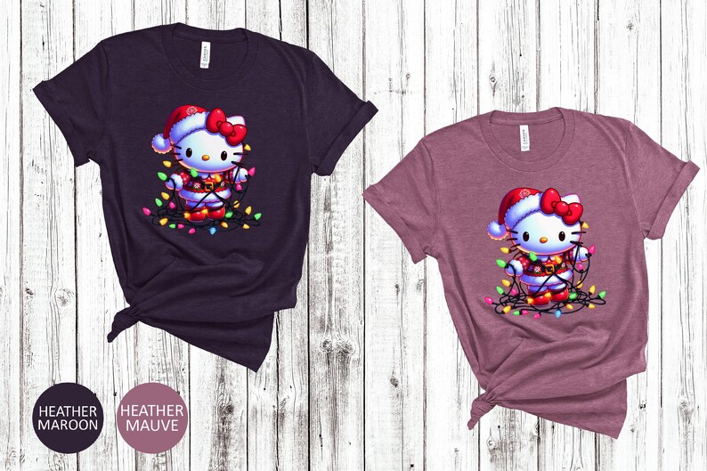 May include: Two t-shirts, one heather maroon and one heather mauve, each featuring a cartoon character wearing a Santa hat and wrapped in Christmas lights. The shirts are displayed on a light wood-grain background.