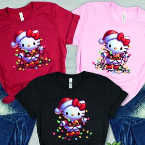 May include: Red, pink, and black t-shirts featuring a graphic of a cartoon cat wearing a Santa hat and wrapped in Christmas lights. The shirts are displayed with blue denim jeans.