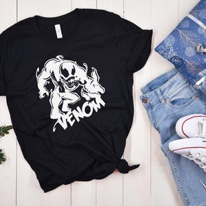 May include: Black t-shirt with a white graphic of Venom, a Marvel Comics character, with the word "Venom" printed below the image.