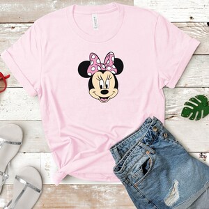 May include: A pink t-shirt with a black and white image of Minnie Mouse's face. Minnie Mouse is wearing a pink bow with white polka dots.
