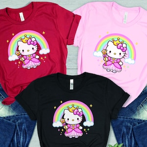 May include: Three short-sleeved t-shirts in red, pink, and black, each featuring a cartoon princess cat design with a rainbow, crown, and wand. The shirts are displayed with blue denim jeans.