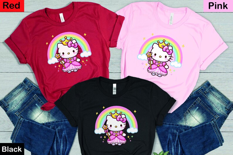 May include: Three short-sleeved t-shirts in red, pink, and black, each featuring a cartoon princess cat design with a rainbow, crown, and wand. The shirts are displayed with blue denim jeans.