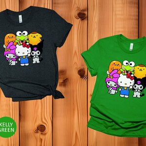 May include: Two t-shirts featuring a graphic print of a group of cartoon characters. The characters include Hello Kitty, My Melody, Keroppi, Badtz-Maru, and a yellow chick. The shirt on the left is heather dark grey and the shirt on the right is kelly green.