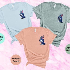 May include: Three t-shirts in different colors: light blue, heather peach, and athletic heather. Each shirt features a small graphic of a blue bunny wearing a hoodie.