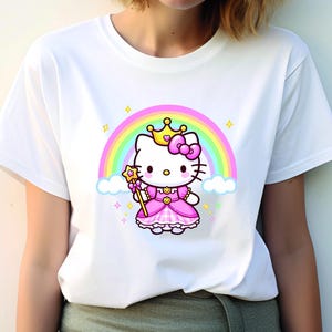 May include: White t-shirt featuring a cartoon princess cat with a crown, bow, and wand, set against a rainbow and clouds. The cat wears a pink dress. The design includes stars and sparkles.