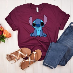 May include: A maroon t-shirt with a blue cartoon character, Stitch, from the Disney movie Lilo & Stitch. The character is sitting down with its arms crossed and has a blue and white design.