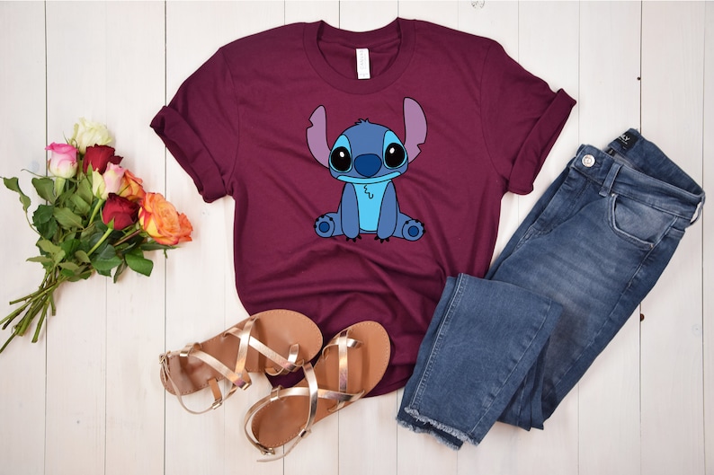 May include: A maroon t-shirt with a blue cartoon character, Stitch, from the Disney movie Lilo & Stitch. The character is sitting down with its arms crossed and has a blue and white design.