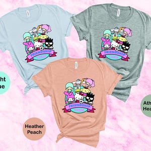 May include: Three t-shirts with a graphic print of cartoon characters. The t-shirts are in light blue, heather peach, and athletic heather colors. The graphic features a group of cartoon characters with the text "Friendship Goals".