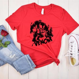 May include: Red t-shirt with a black graphic of Venom, a Marvel Comics character, and the word "VENOM" printed below. The shirt is laid out on a white wooden surface with a pair of white sneakers and blue jeans with a red rose.