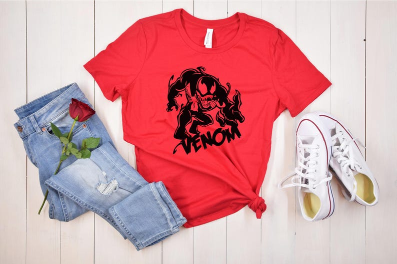 May include: Red t-shirt with a black graphic of Venom, a Marvel Comics character, and the word "VENOM" printed below. The shirt is laid out on a white wooden surface with a pair of white sneakers and blue jeans with a red rose.
