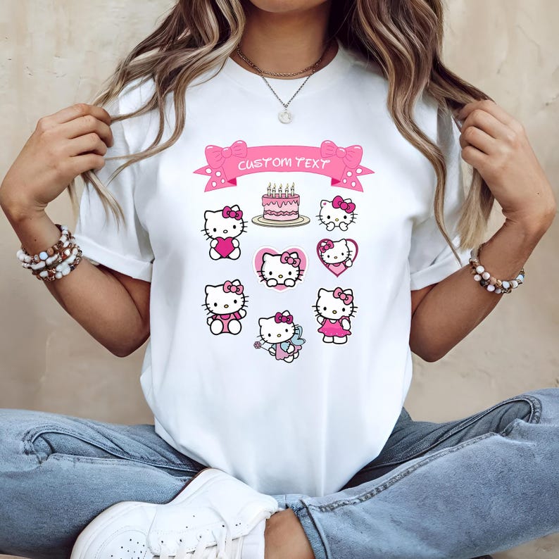 May include: White t-shirt featuring multiple Hello Kitty designs in pink and red. A pink banner with the text "CUSTOM TEXT" is above a cake with lit candles. The shirt has a crew neck and short sleeves.