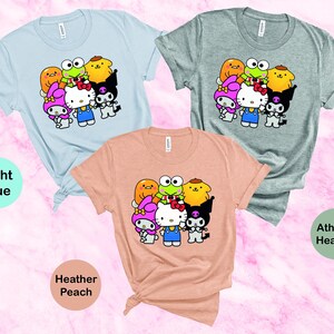 May include: Three t-shirts featuring a graphic print of cartoon characters. The t-shirts are in light blue, heather peach and athletic heather colours. The graphic features Hello Kitty and other cartoon characters.