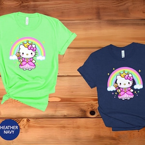 May include: Two short-sleeved t-shirts, one mint green and one navy blue, featuring a cartoon princess cat design. The cat wears a crown and holds a wand in front of a rainbow. The shirts are displayed on a wooden surface.