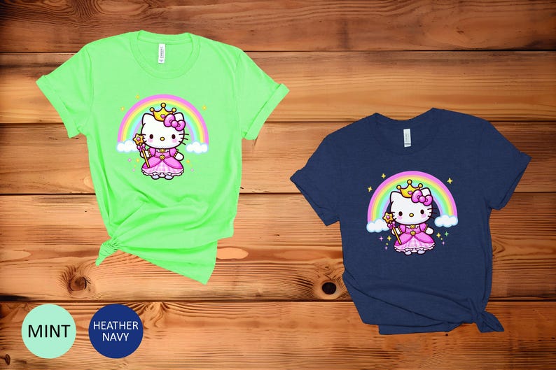 May include: Two short-sleeved t-shirts, one mint green and one navy blue, featuring a cartoon princess cat design. The cat wears a crown and holds a wand in front of a rainbow. The shirts are displayed on a wooden surface.