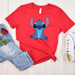 May include: Red t-shirt with a blue cartoon Stitch character graphic. The shirt is laid out on a white wooden surface with blue jeans and white sneakers.