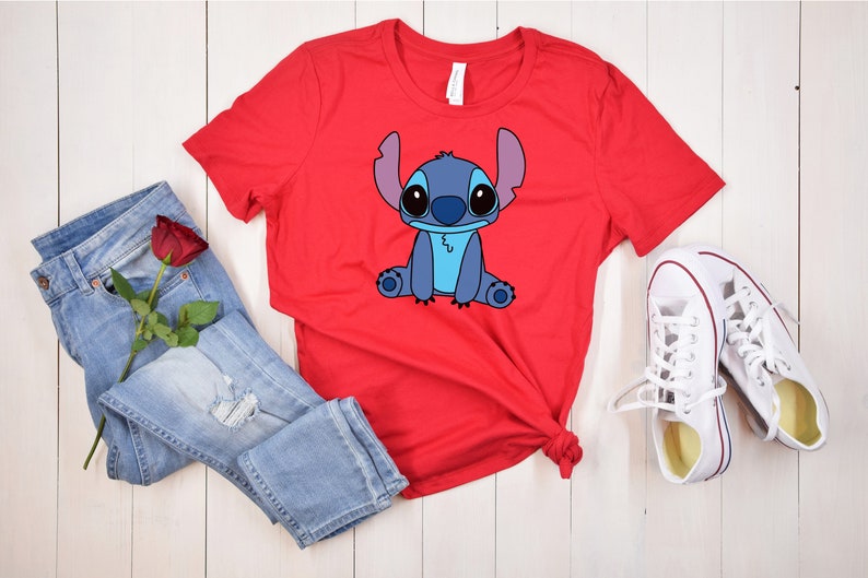 May include: Red t-shirt with a blue cartoon Stitch character graphic. The shirt is laid out on a white wooden surface with blue jeans and white sneakers.