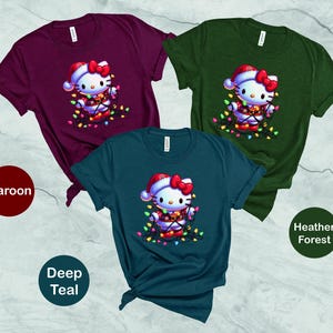 May include: Three t-shirts in maroon, deep teal, and heather forest green. Each shirt features a graphic of a cartoon cat wearing a Santa hat and wrapped in Christmas lights. The shirts are short-sleeved.