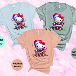 May include: Three short-sleeved t-shirts in light blue, athletic heather, and heather peach. Each shirt features a graphic of a cartoon cat wearing a Santa hat and wrapped in Christmas lights. The shirts are knotted at the bottom.
