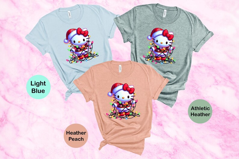 May include: Three short-sleeved t-shirts in light blue, athletic heather, and heather peach. Each shirt features a graphic of a cartoon cat wearing a Santa hat and wrapped in Christmas lights. The shirts are knotted at the bottom.
