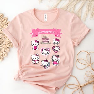 May include: Pink t-shirt featuring multiple Hello Kitty designs with bows, hearts, and a birthday cake. A pink banner with the text "CUSTOM TEXT" is above the designs. The shirt is made of a soft material.
