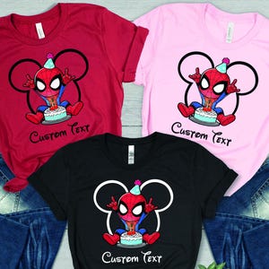 May include: Red, pink, and black t-shirts featuring a cartoon Spider-Man design. The character wears a party hat and sits in front of a cake. The text "Custom Text" is below the image. Blue jeans are also in the image.