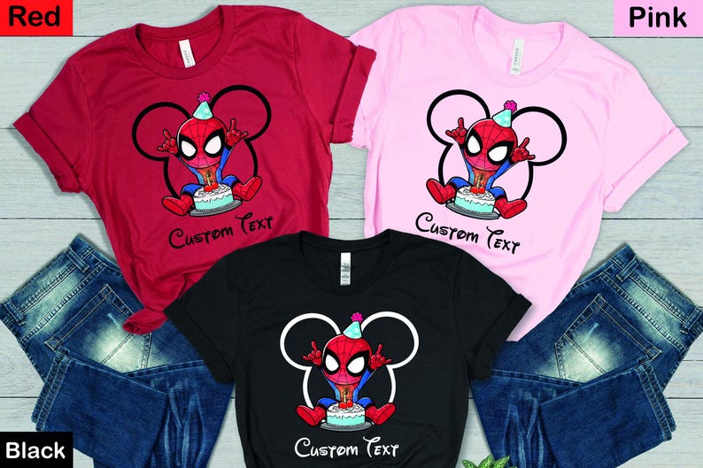 May include: Red, pink, and black t-shirts featuring a cartoon Spider-Man design. The character wears a party hat and sits in front of a cake. The text "Custom Text" is below the image. Blue jeans are also in the image.