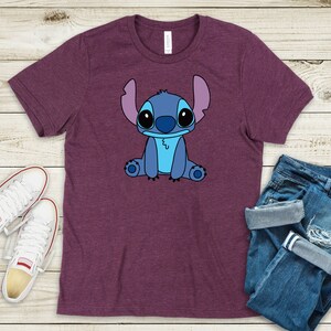 May include: A purple t-shirt with a cartoon image of Stitch from Lilo & Stitch. Stitch is blue with large eyes and is sitting down.