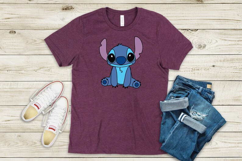May include: A purple t-shirt with a cartoon image of Stitch from Lilo & Stitch. Stitch is blue with large eyes and is sitting down.