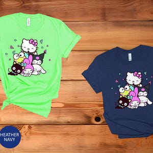 Pu&ograve; includere: Due t-shirt con una stampa grafica di Hello Kitty e i suoi amici. La maglietta a sinistra &egrave; di colore verde brillante e la maglietta a destra &egrave; di colore blu scuro.