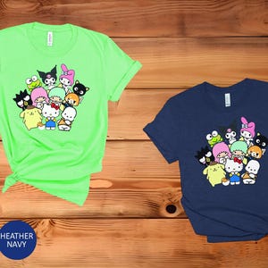 May include: Two t-shirts with a graphic print of a group of cartoon characters. The t-shirt on the left is a bright green color and the t-shirt on the right is a dark blue color.