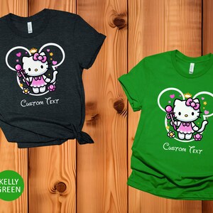 May include: Two t-shirts with a cartoon image of Hello Kitty dressed as a princess with a wand and a crown. The image is inside a Mickey Mouse ear shape. The text "Custom Text" is below the image. One shirt is heather dark grey and the other is kelly green.