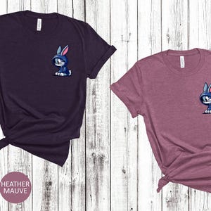 May include: Two t-shirts with a cartoon bunny wearing a blue hoodie on the front pocket. The shirt on the left is heather maroon and the shirt on the right is heather mauve.