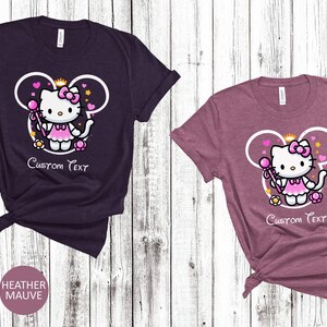 May include: Two t-shirts with a pink Hello Kitty character wearing a crown and holding a wand. The character is inside a white outline of Mickey Mouse ears. The text "Custom Text" is below the character. The t-shirts are heather maroon and heather mauve.