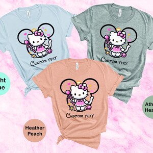 May include: Three t-shirts with a cartoon image of Hello Kitty wearing a pink dress and holding a wand. The t-shirts are in light blue, heather peach, and athletic heather colors. The text "CUSTOM TEXT" is printed below the image on each shirt.