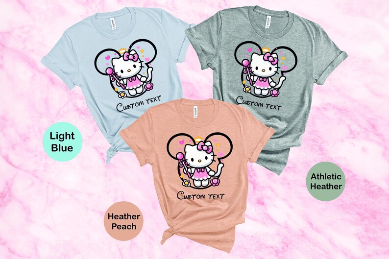 May include: Three t-shirts with a cartoon image of Hello Kitty wearing a pink dress and holding a wand. The t-shirts are in light blue, heather peach, and athletic heather colors. The text "CUSTOM TEXT" is printed below the image on each shirt.