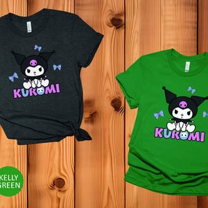 Puede incluir: Dos camisetas, una gris oscuro jaspeado y otra verde Kelly, cada una con un dise&ntilde;o de personaje de dibujos animados. El dise&ntilde;o incluye un personaje negro con una calavera, lazos azules y la palabra "KUROMI" en rosa y morado.