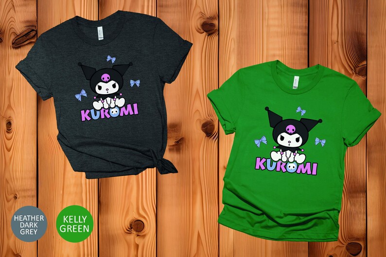 Puede incluir: Dos camisetas, una gris oscuro jaspeado y otra verde Kelly, cada una con un dise&ntilde;o de personaje de dibujos animados. El dise&ntilde;o incluye un personaje negro con una calavera, lazos azules y la palabra "KUROMI" en rosa y morado.