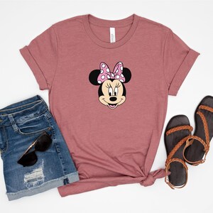 May include: A mauve t-shirt with a cartoon image of Minnie Mouse wearing a pink bow with white polka dots. The t-shirt is paired with denim shorts and brown sandals.