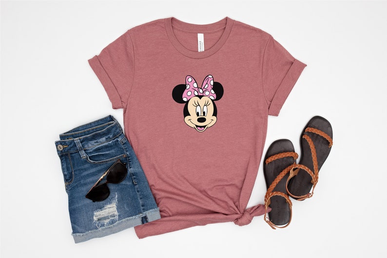 May include: A mauve t-shirt with a cartoon image of Minnie Mouse wearing a pink bow with white polka dots. The t-shirt is paired with denim shorts and brown sandals.