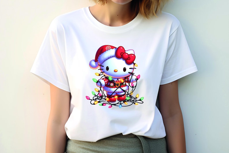 May include: White t-shirt featuring a festive Hello Kitty design. The cartoon character is dressed in a Santa hat and suit, adorned with a red bow and wrapped in colorful Christmas lights. A fun and cheerful holiday-themed graphic.